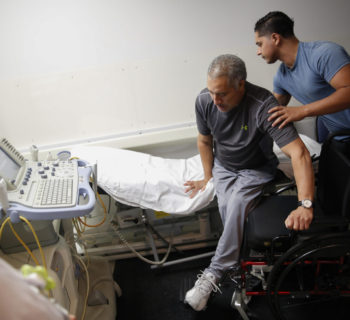 Joel Ramirez is helped onto a bed to receive an ultrasound with the aid of Francisco Guardado, his home health care giver, during a visit to the hospital in Rialto, Calif.